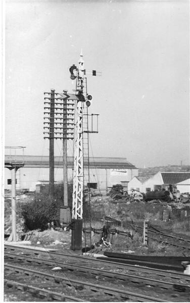 A wooden latice post signal carrying a home and distant signal arm on opposite side of the post, viewd from a passing train across the goods lines with Derby Corporation Sidings in the background